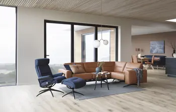 Modernes Wohnzimmer mit einem Stressless Stella Sofa in Paloma Copper Leder und einem blauen Stressless Tokyo Sessel mit passendem Fußhocker. Große Fenster bieten einen Blick auf die Landschaft, und die Einrichtung ist minimalistisch gehalten.